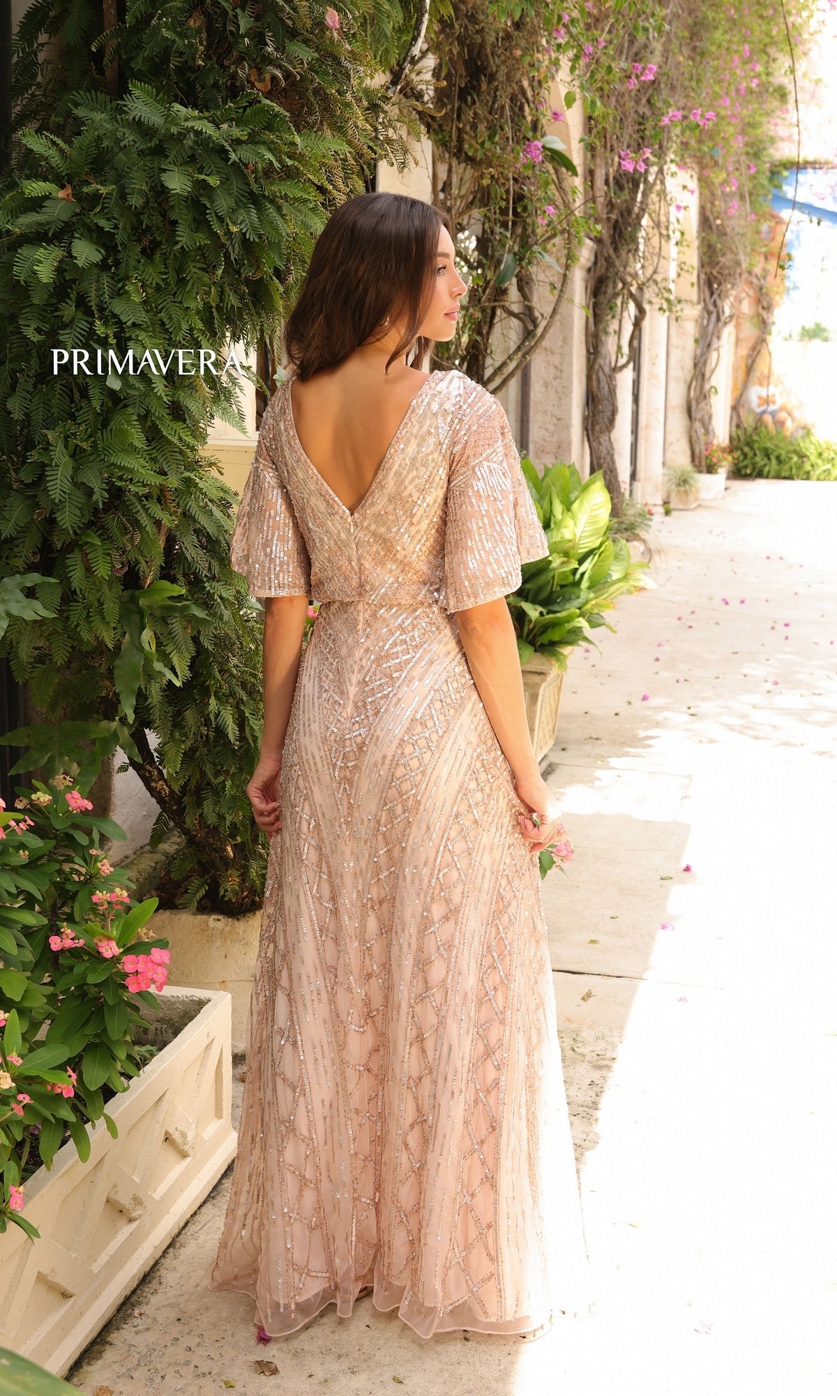Woman in the Primavera 13119 Sleeved Long Sequin Formal Dress, a shimmering light pink gown with short flutter sleeves, stands on a sunlit walkway surrounded by greenery and flowers.